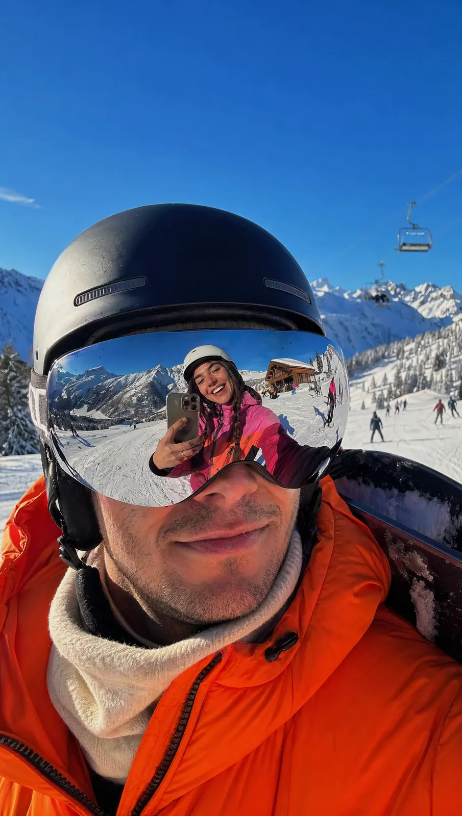 A skier in an orange jacket smiles under a black helmet; his mirrored goggles reflect a woman taking a selfie on the slopes